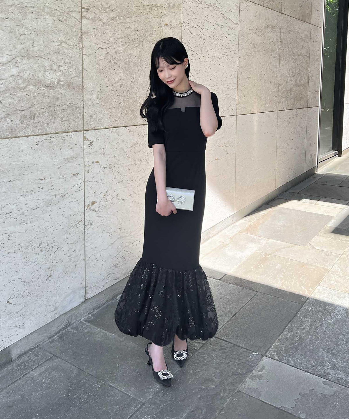 首元のビジューと透け感のあるデコルテ、裾のラメ入りバルーンヘムが華やかな黒のマーメイドオケージョンドレスを着用した女性の立ち姿。