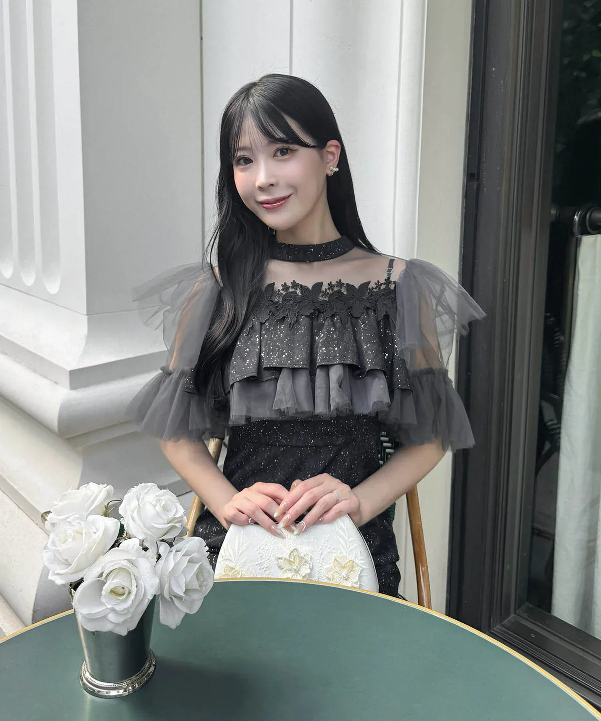 黒地にラメとチュールフリルを重ねたエンブロイダリーワンピースを着てテラス席に座る女性。結婚式やお呼ばれにおすすめの上品なデザイン。