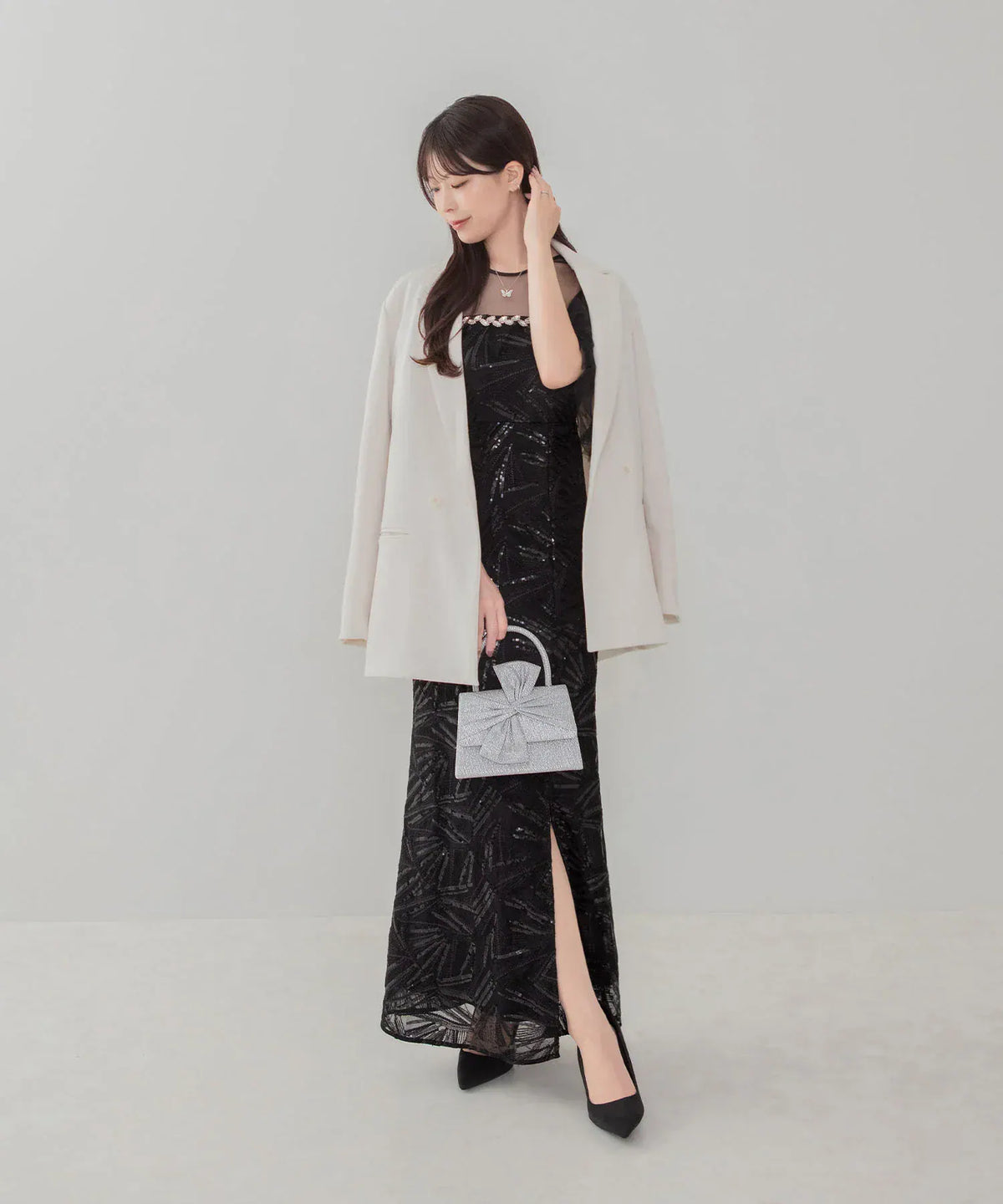 黒のスリット入り総柄ロングドレスに白いジャケットを羽織り手元に持った、シルバーに輝くビッグリボン付きパーティーバッグのコーディネート。