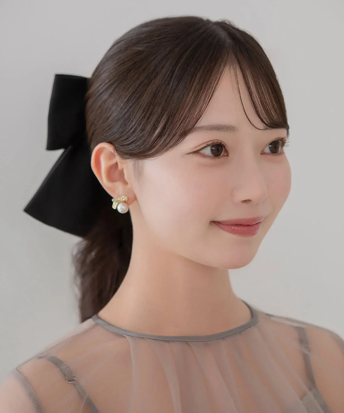 まとめ髪の後ろ姿に映える黒のビッグリボンメタルプレートバレッタをつけた女性のサイドアップ。シアーなドレスとパールイヤリングを合わせた華やかなスタイリング。