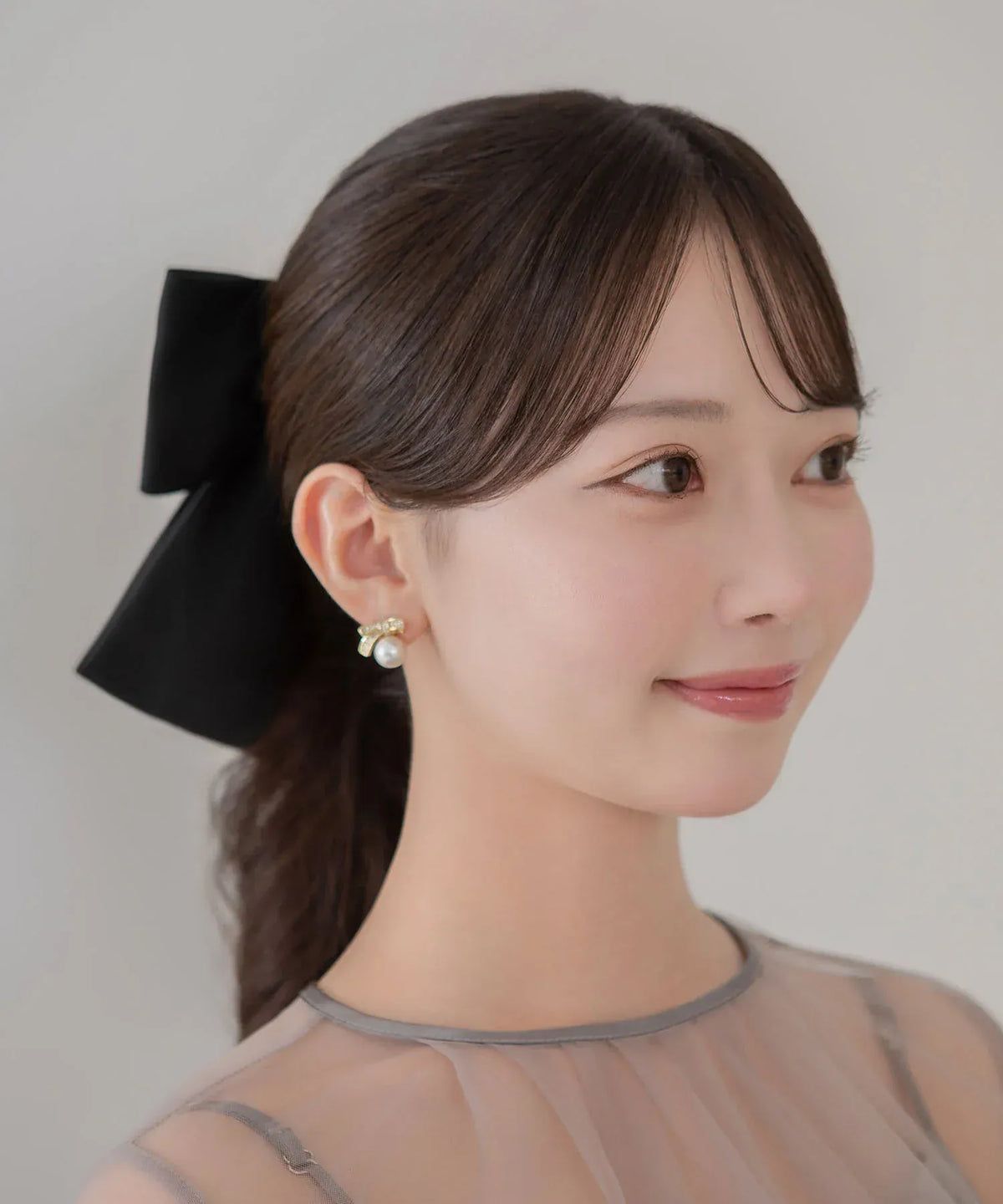 透け感のあるドレスを着た女性がポニーテールに黒のビッグリボンメタルプレートバレッタとパールイヤリングを合わせた横顔アップ。