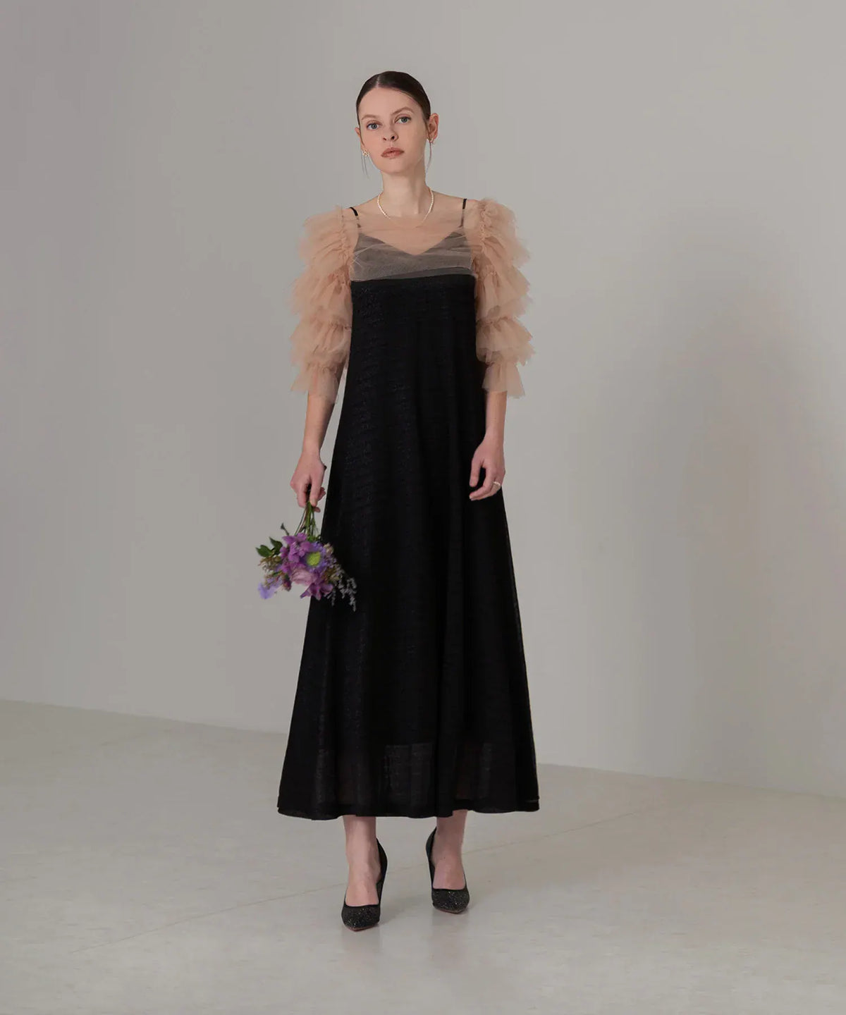 ベージュのチュールフリルボレロに黒のキャミソールワンピースを重ね、花束を持って上品に佇む女性のオケージョンドレス姿。