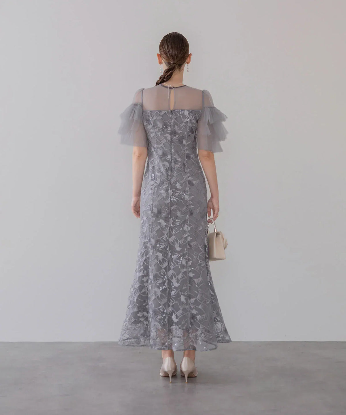 透け感のあるシアーチュール袖とバックファスナーが際立つ、上品なグレーの総柄刺繍マーメイドロングワンピースの後ろ姿。