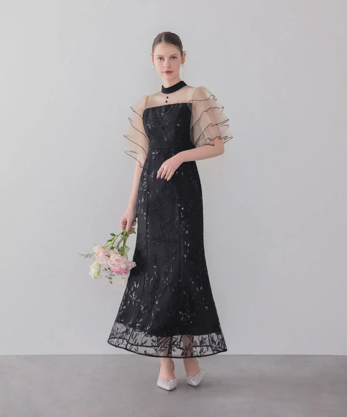 ベージュの段フリル袖と黒のスパンコール刺繍が華やかな、ハイネックのマーメイドオケージョンドレスの全身スタイル。
