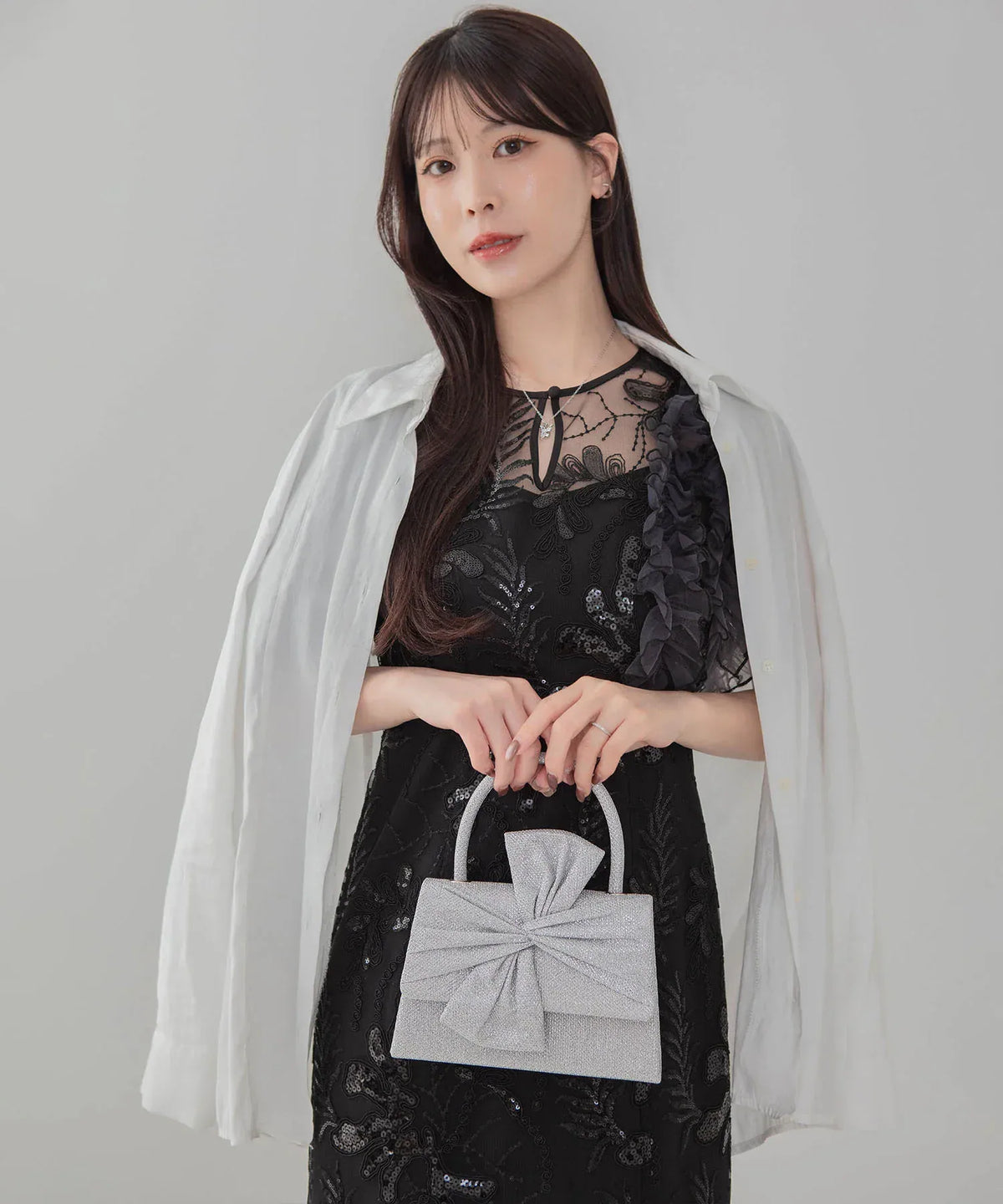 黒の刺繍レースワンピースと白シャツに合わせた、シルバーの立体的な大ぶりリボンが華やかなグリッターハンドバッグの手持ちスタイル。