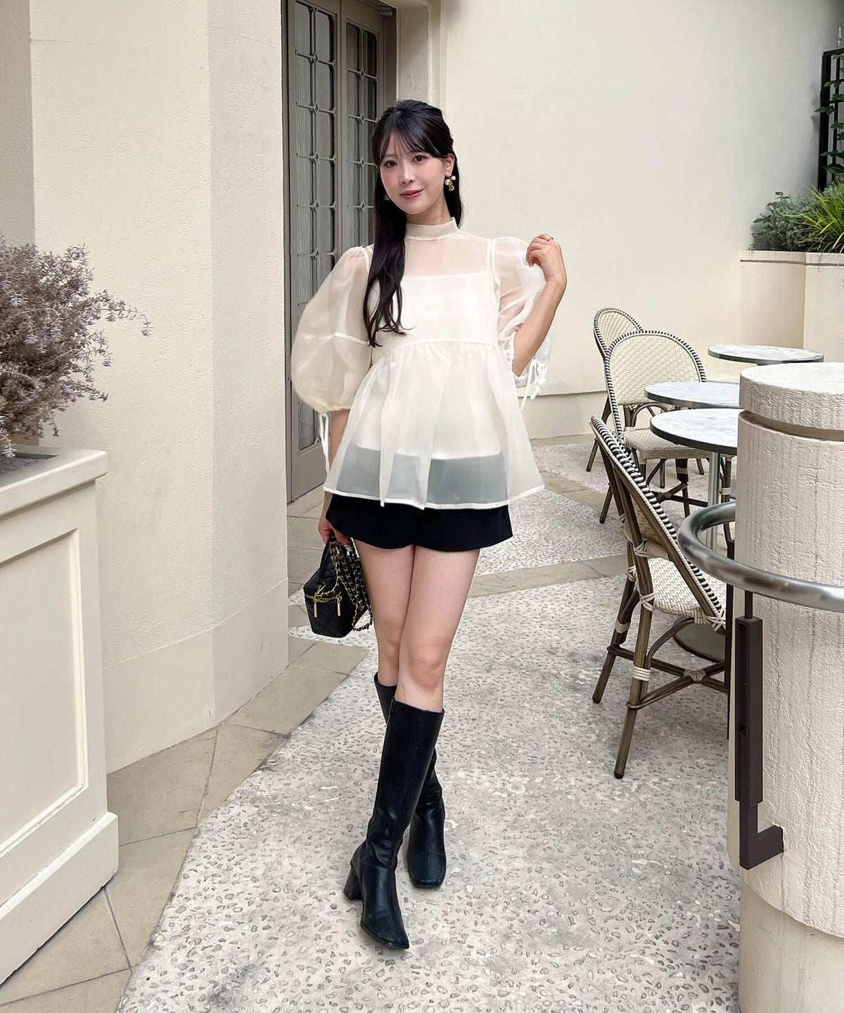 ふんわりとしたボリューム袖に黒のショートパンツとロングブーツを合わせた、透け感のある白のシアーオーガンジーハイネックブラウスの着こなし。