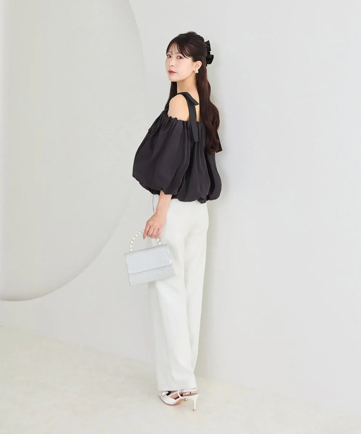 白のワイドパンツに合わせた、ふんわり広がる黒のボリューム袖と肩リボンが印象的な2WAYショルダーリボンバルーンオフショルトップスの横姿
