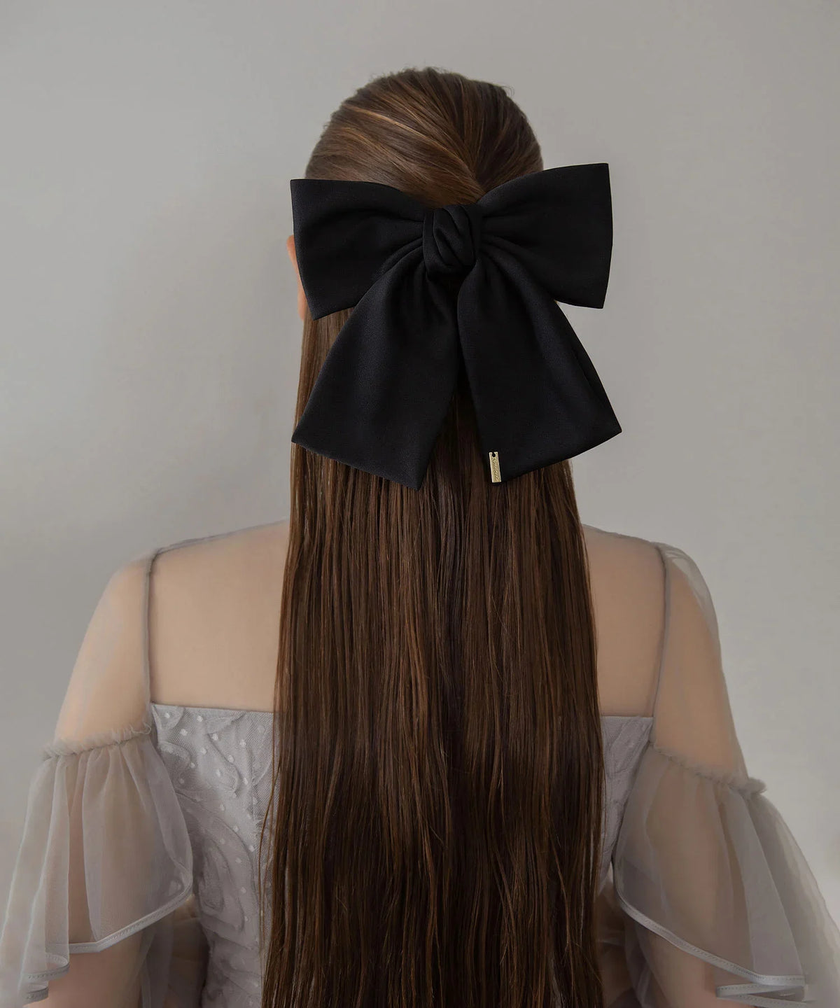 ハーフアップのロングヘアに黒のビッグリボンメタルプレートバレッタを着け、透け感のあるドレスを着た女性の後ろ姿。